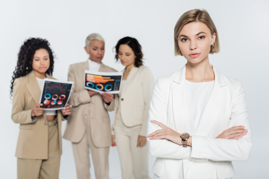 blonde businesswoman looking at camera near blurred interracial colleagues with papers isolated on