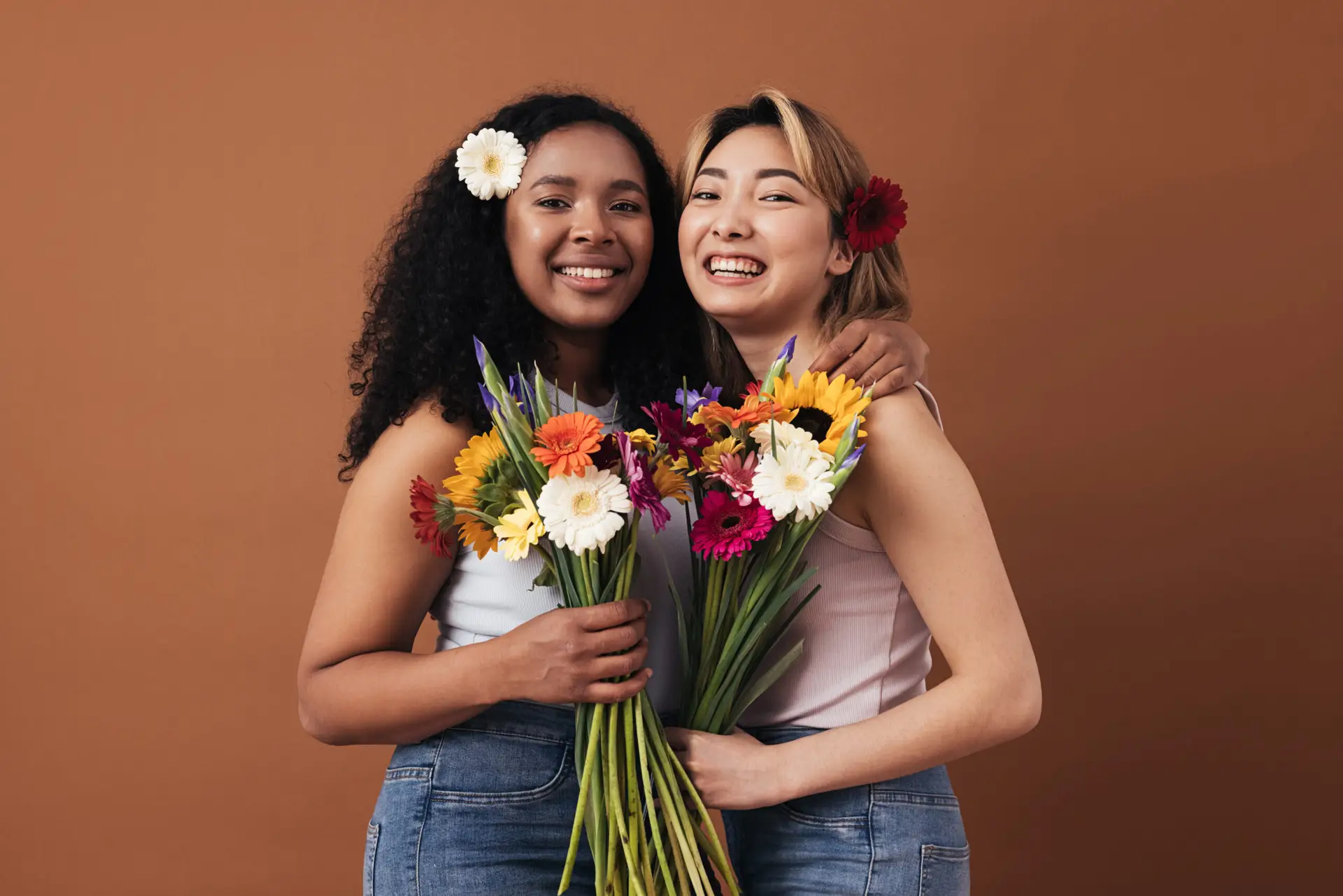 two young women of different races with bouquets and flowers in their hair posing against a brown background